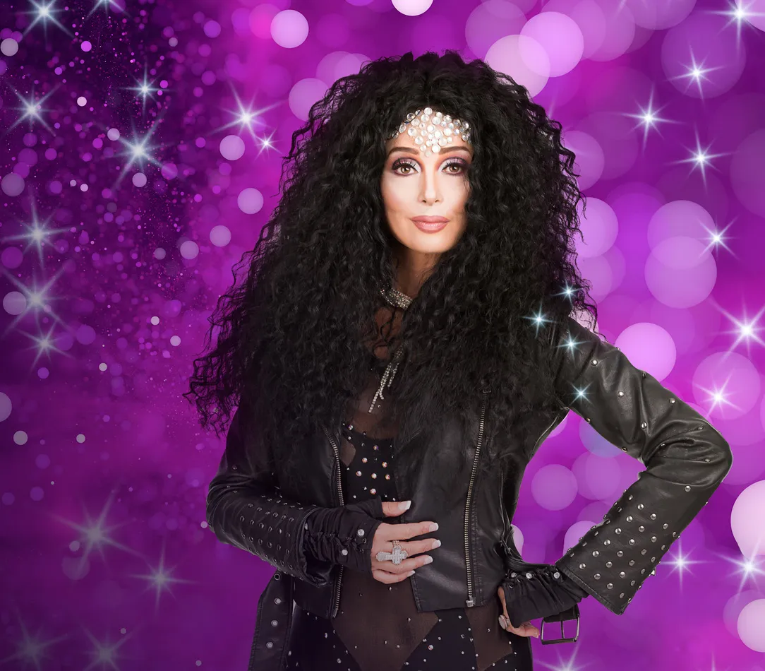 A performer dressed as Cher singing on stage with a band.